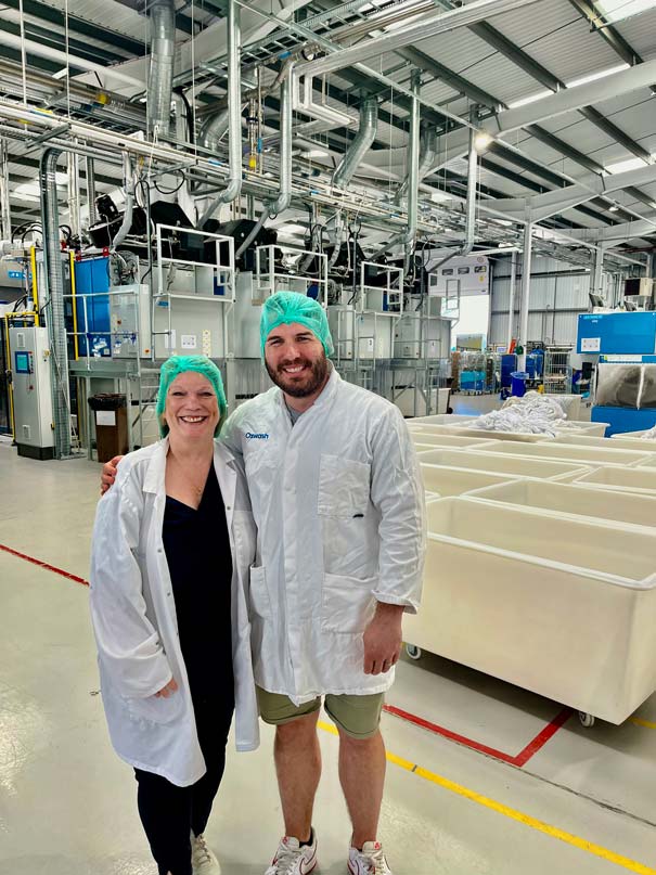 a man and a woman stand in a linen washing warehouse wearing green hair nets and white lab coats, smiling at the camera