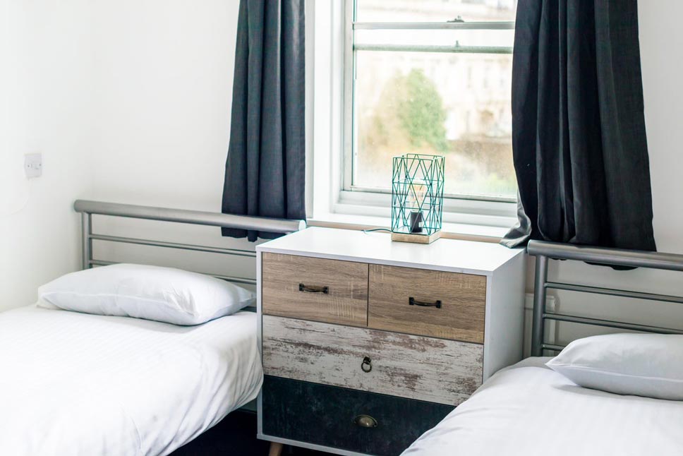 two twin beds with a dresser in the middle of them and a window behind it with Georgian buildings in the background
