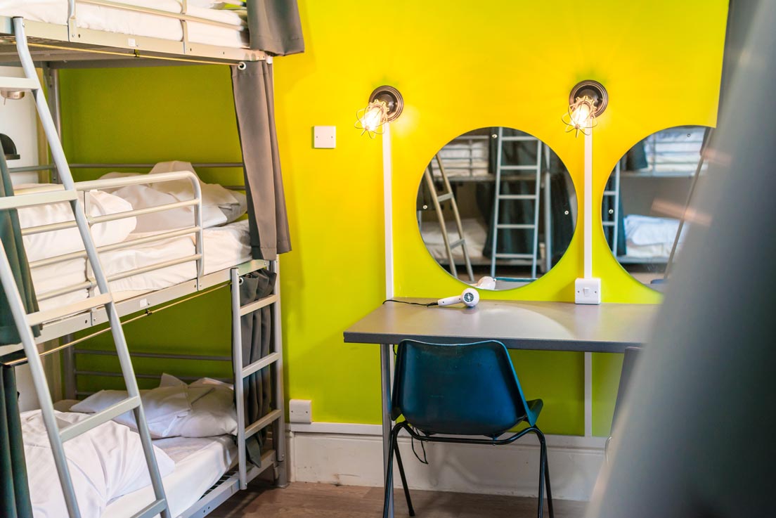 a hostel dorm room with beds on the left and a table against a bright green wall with mirrors and a hair dryer