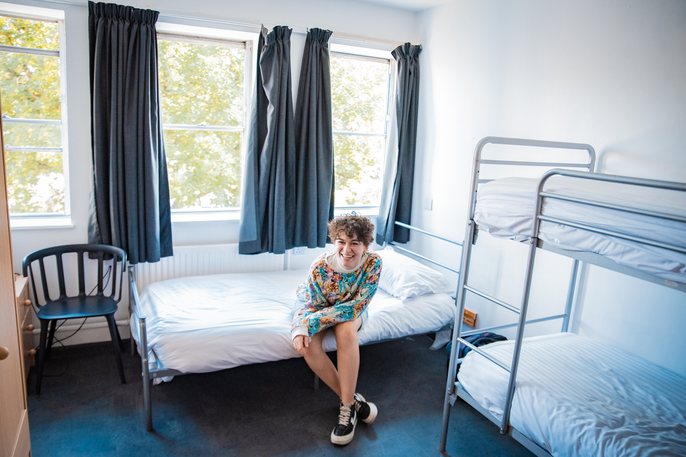 a young woman with short brown curly hair sits on a single bed smiling in a hostel room with two other single beds